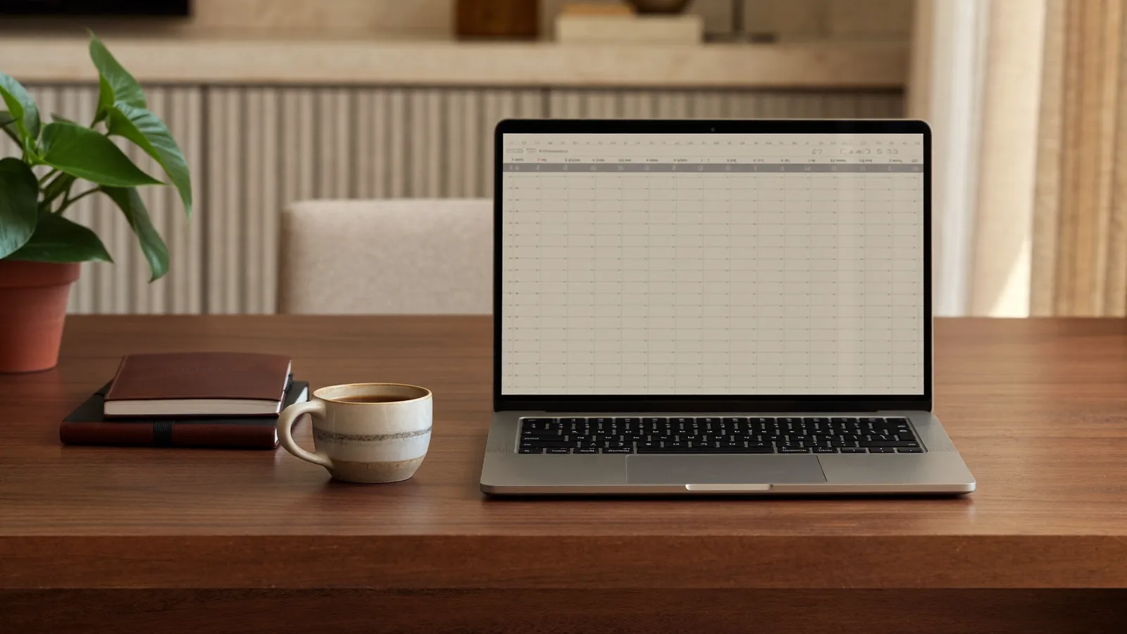 Una laptop sobre un escritorio de madera con una hoja de cálculo vacía en la pantalla, junto a una taza de café y una libreta cerrada.