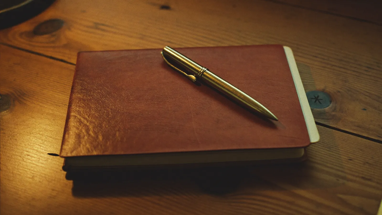 Una agenda de piel cerrada con una pluma encima, sobre una mesa de madera clara.